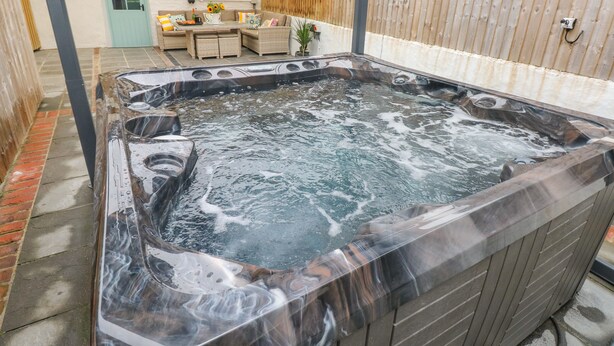 A hot tub with a wooden exterior, a patio area with a table and chairs, and a wooden fence in the background. - Y Beudy (Crymych)