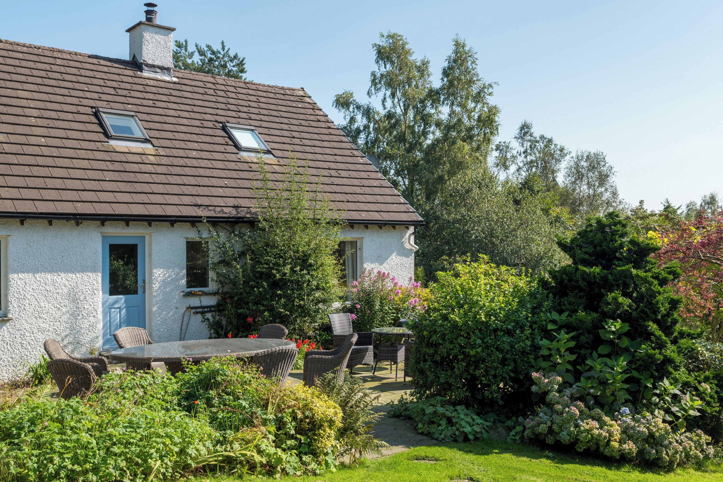 A house with a tiled roof, a blue door, and a garden with various plants and a table with chairs.