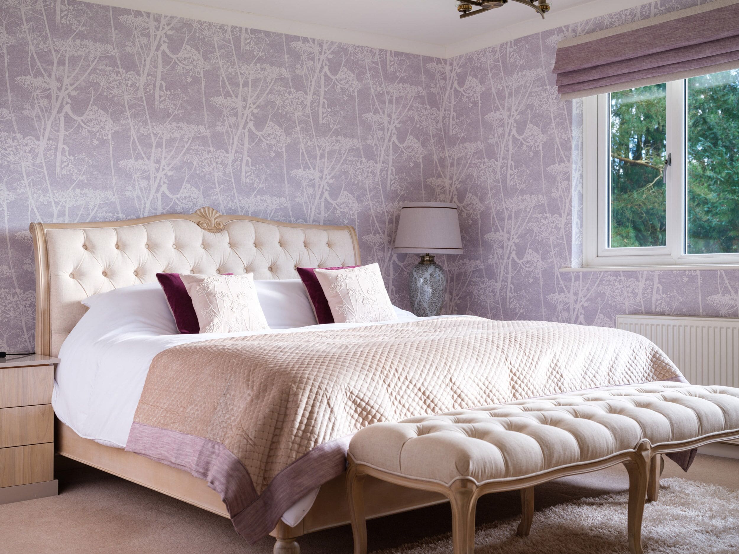 A bedroom with a tufted headboard, a large bed with a quilted bedspread, a bedside table, a lamp, and a window with a view of greenery.
