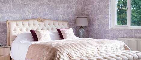 A bedroom with a tufted headboard, a large bed with a quilted bedspread, a bedside table, a lamp, and a window with a view of greenery.