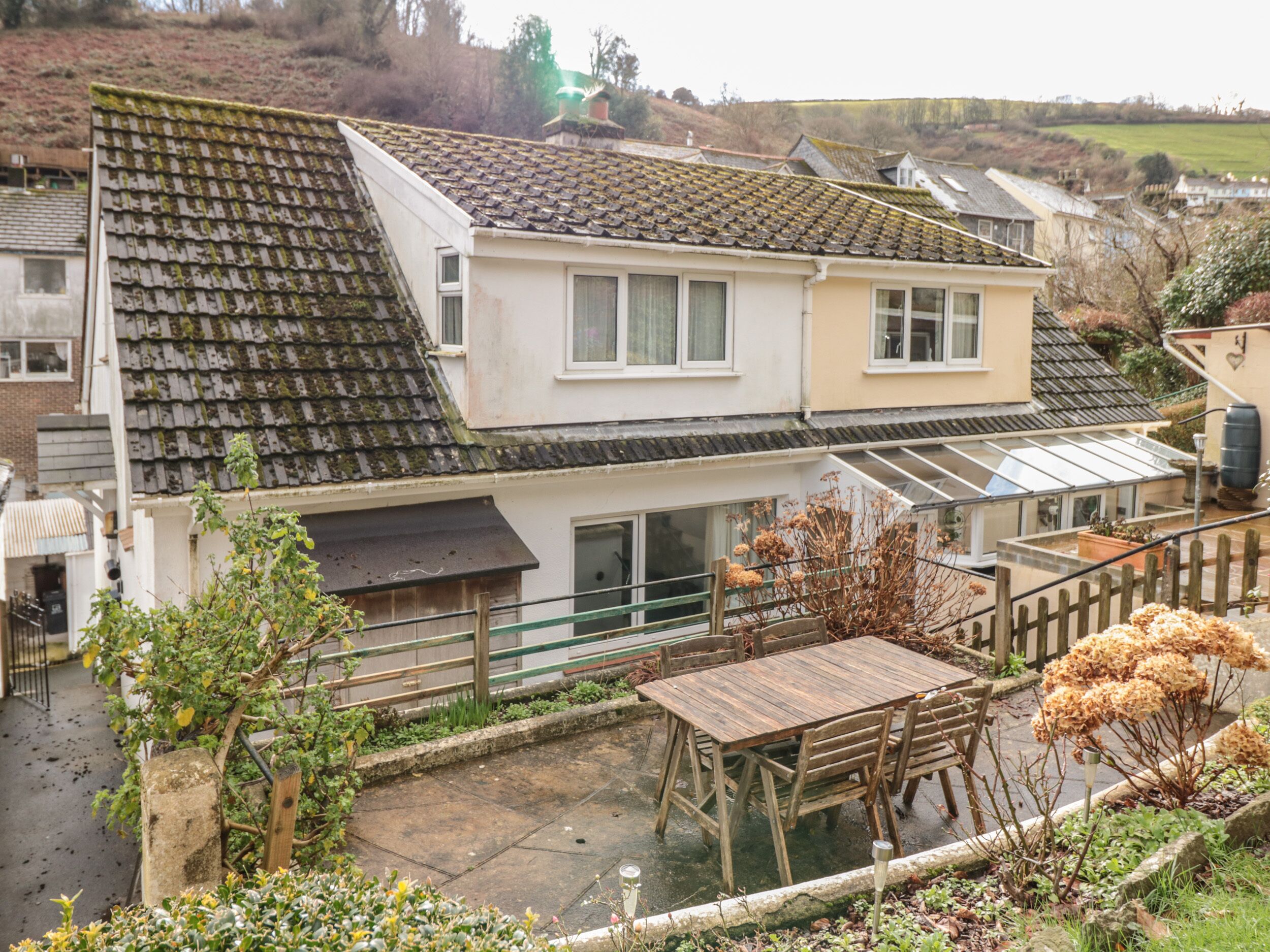 A two-story house with a sloped roof, a patio with a wooden table and chairs, and a garden area with various plants.