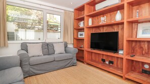 A living room with a grey sofa, a wooden entertainment unit with a flat-screen TV, and large windows with curtains. - George Cottage (Dartmouth)