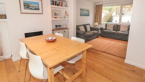 A modern living room with a wooden dining table, white chairs, a grey sofa, and a bookshelf with books and decorative items. - George Cottage (Dartmouth)