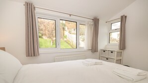 A bedroom with a large bed, a window with curtains, a radiator, and a small white dresser with baskets. - George Cottage (Dartmouth)