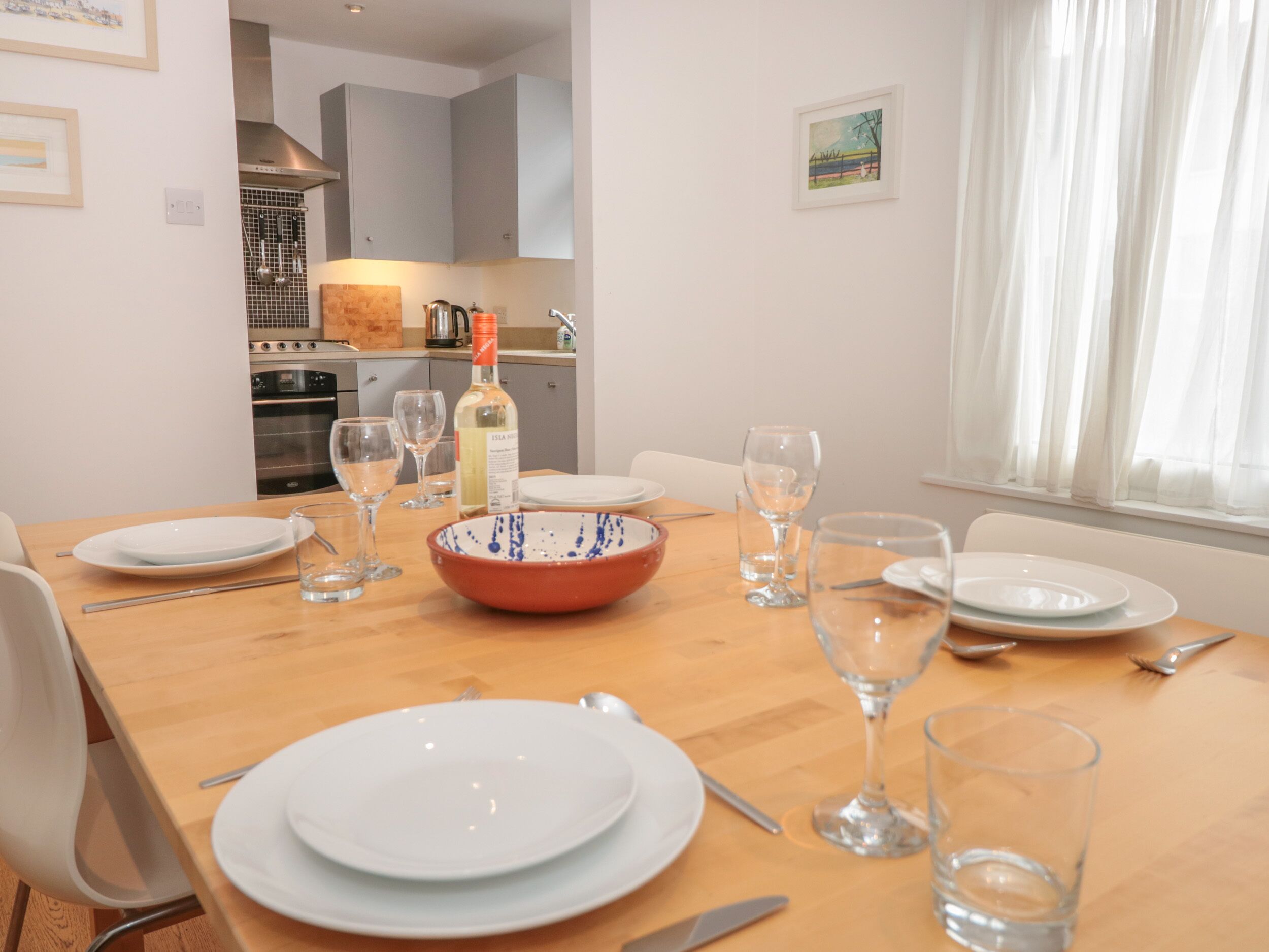 Una mesa de comedor con platos, vasos y una botella, en una habitación con cocina al fondo.