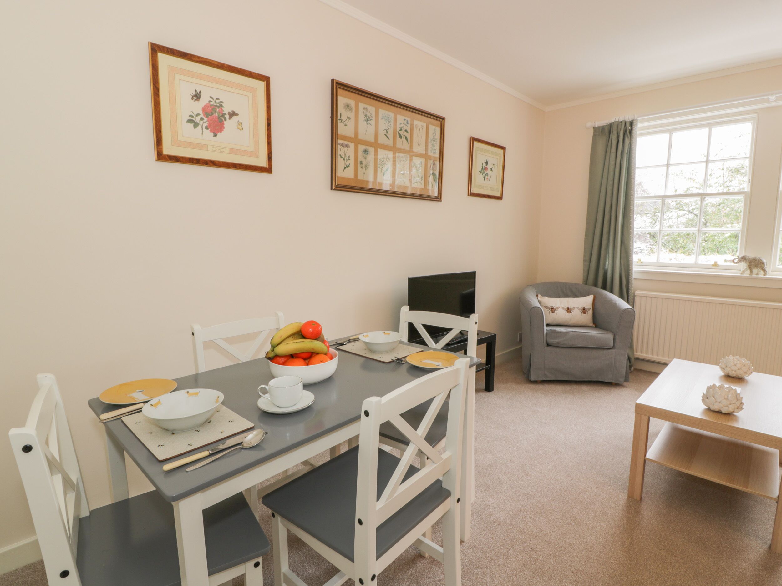 A dining area with a table set for two, a grey sofa, and framed botanical prints on the wall.