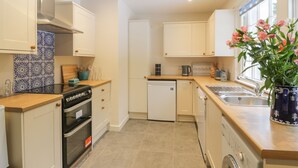 A modern kitchen with white cabinets, a stove, oven, dishwasher, and a sink. There is a window with a view of the outside. - Maisie's Cottage (Dunblane)
