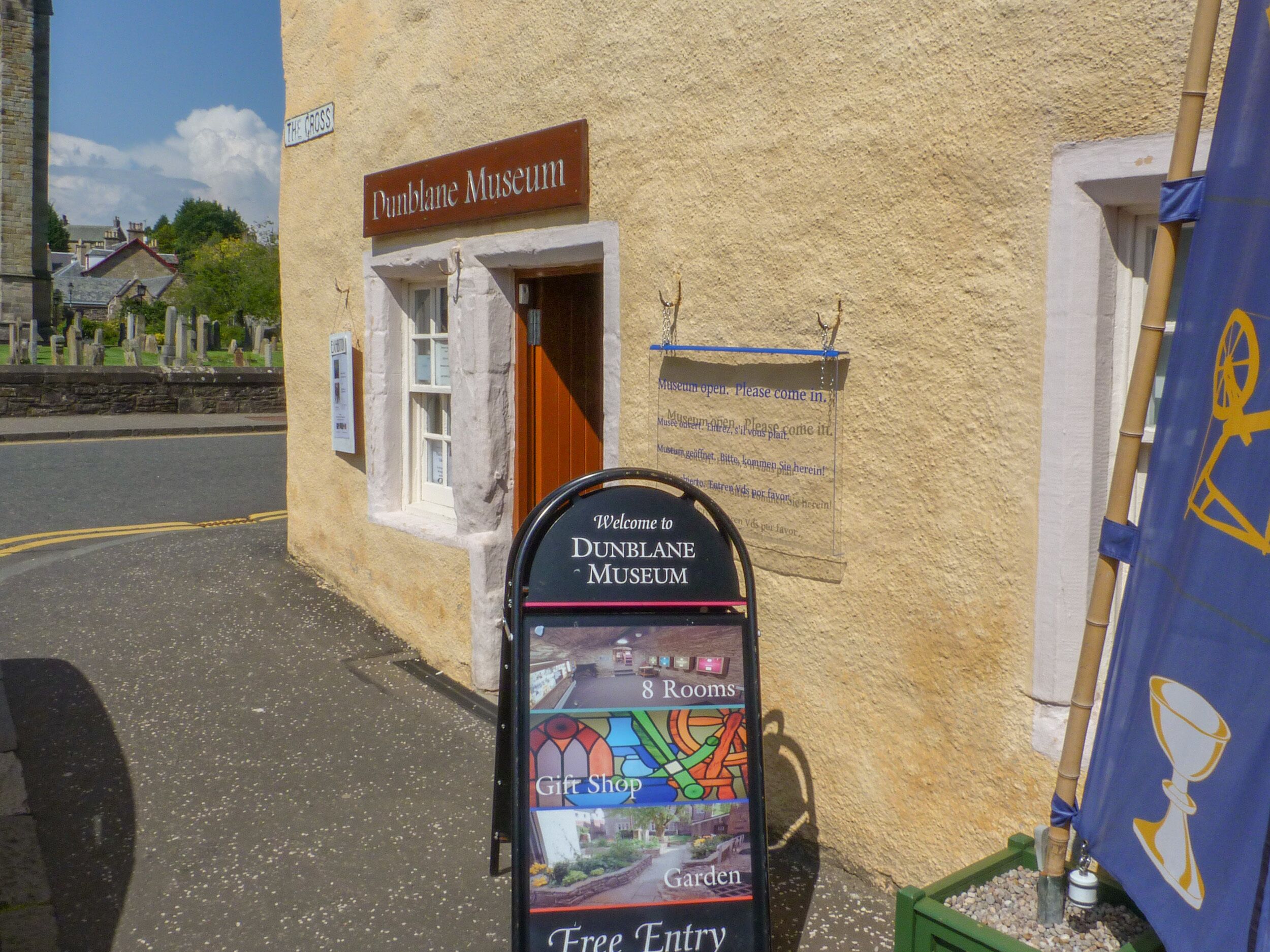 A sign for the Dunblane Museum with a welcome message and information about 8 rooms, a gift shop, and a garden.