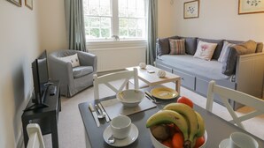 A cozy living room with a grey sofa, a dining table with a bowl of fruit, and a window with curtains. - Maisie's Cottage (Dunblane)