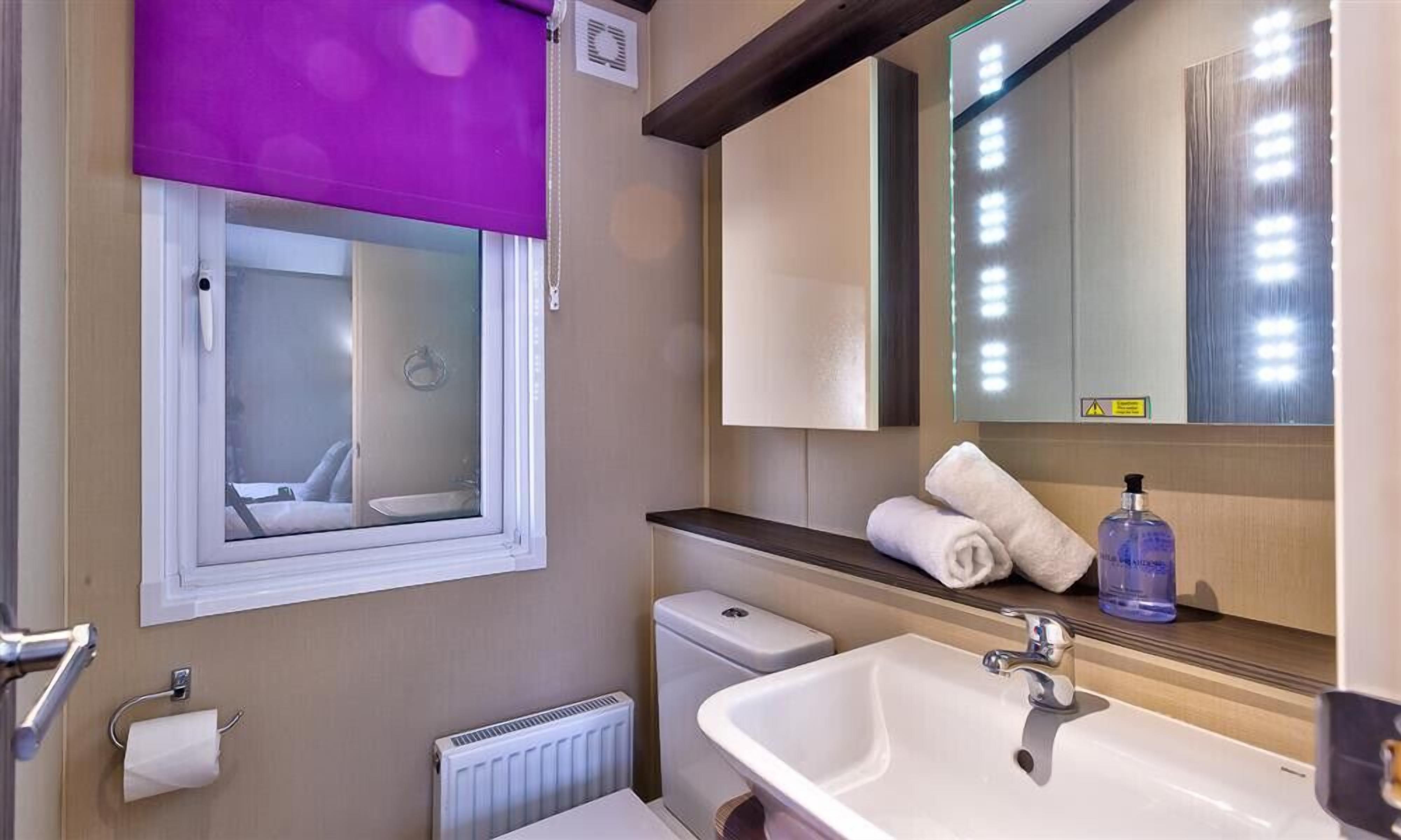 A bathroom with a white sink, a mirror, a window with a purple blind, a towel rack, and a toilet.