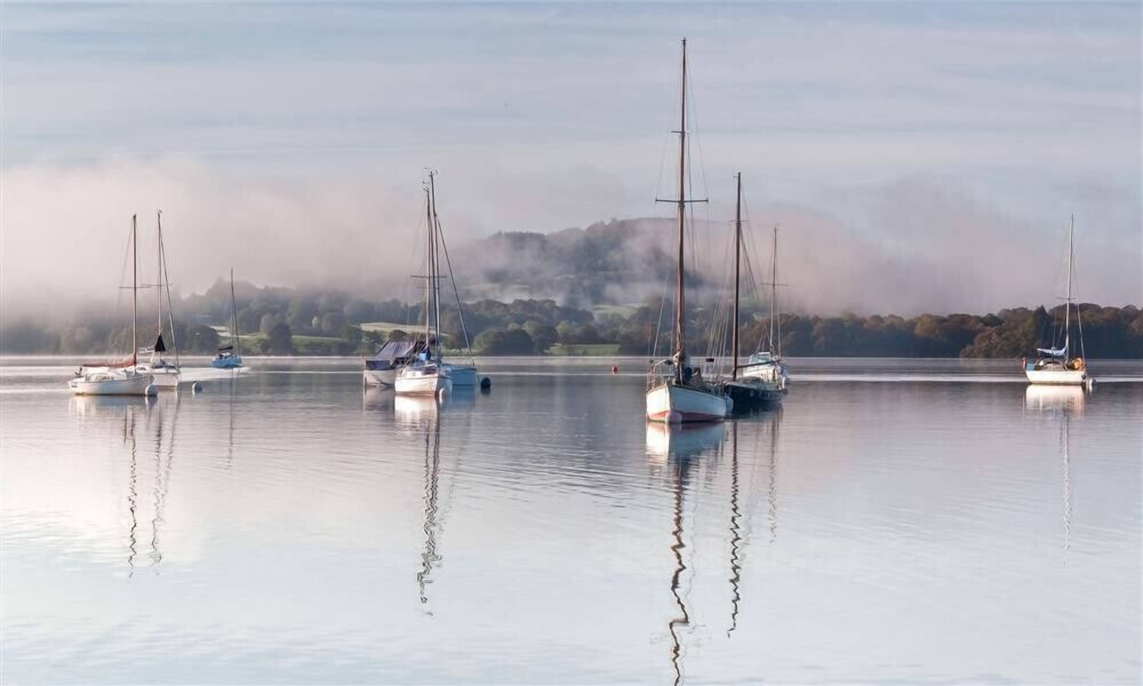 Un llac tranquil amb diversos velers amarrats, envoltat de turons brumosos.