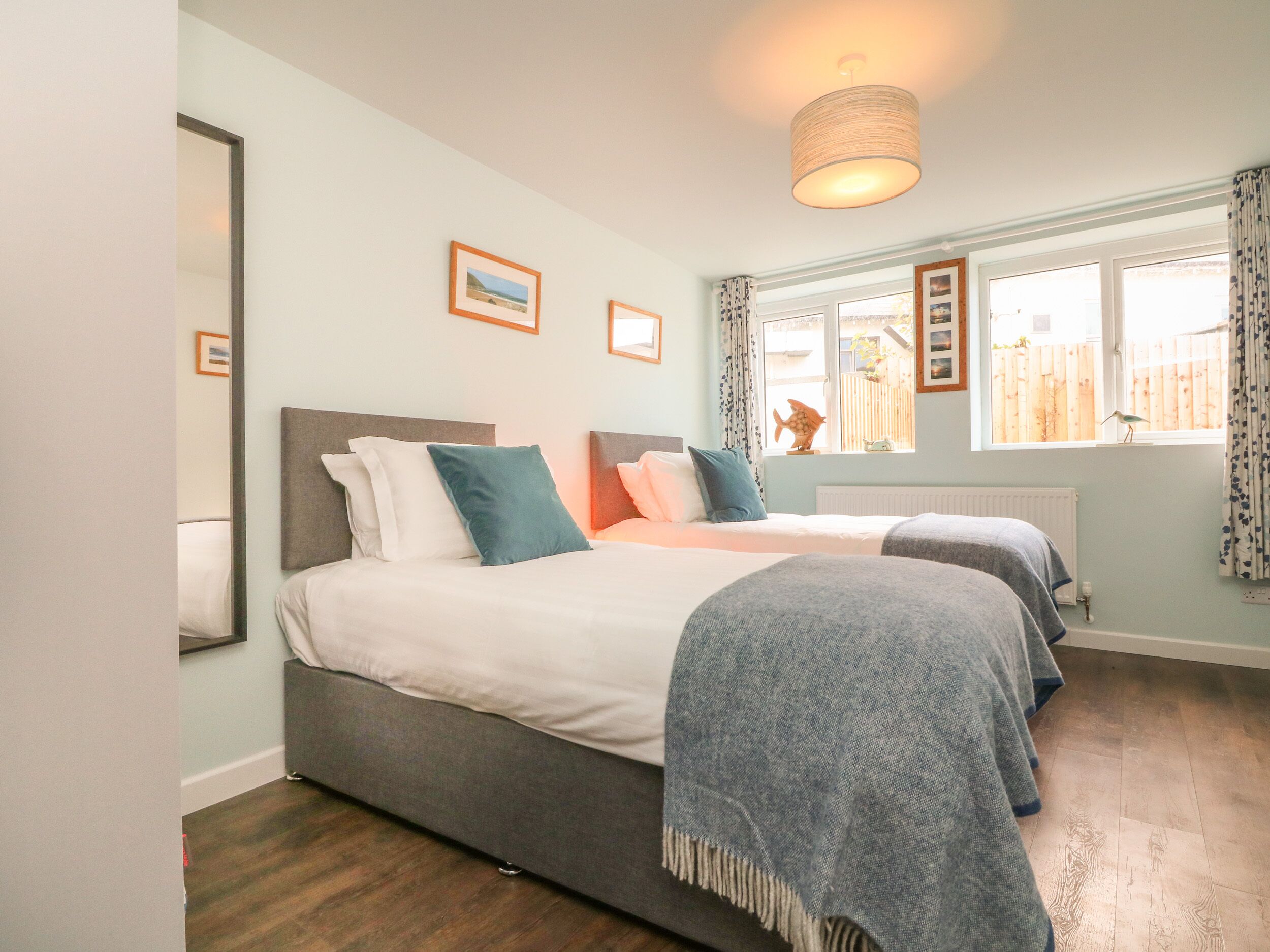 A bedroom with two beds, a large window with curtains, a hanging light fixture, and framed pictures on the wall.