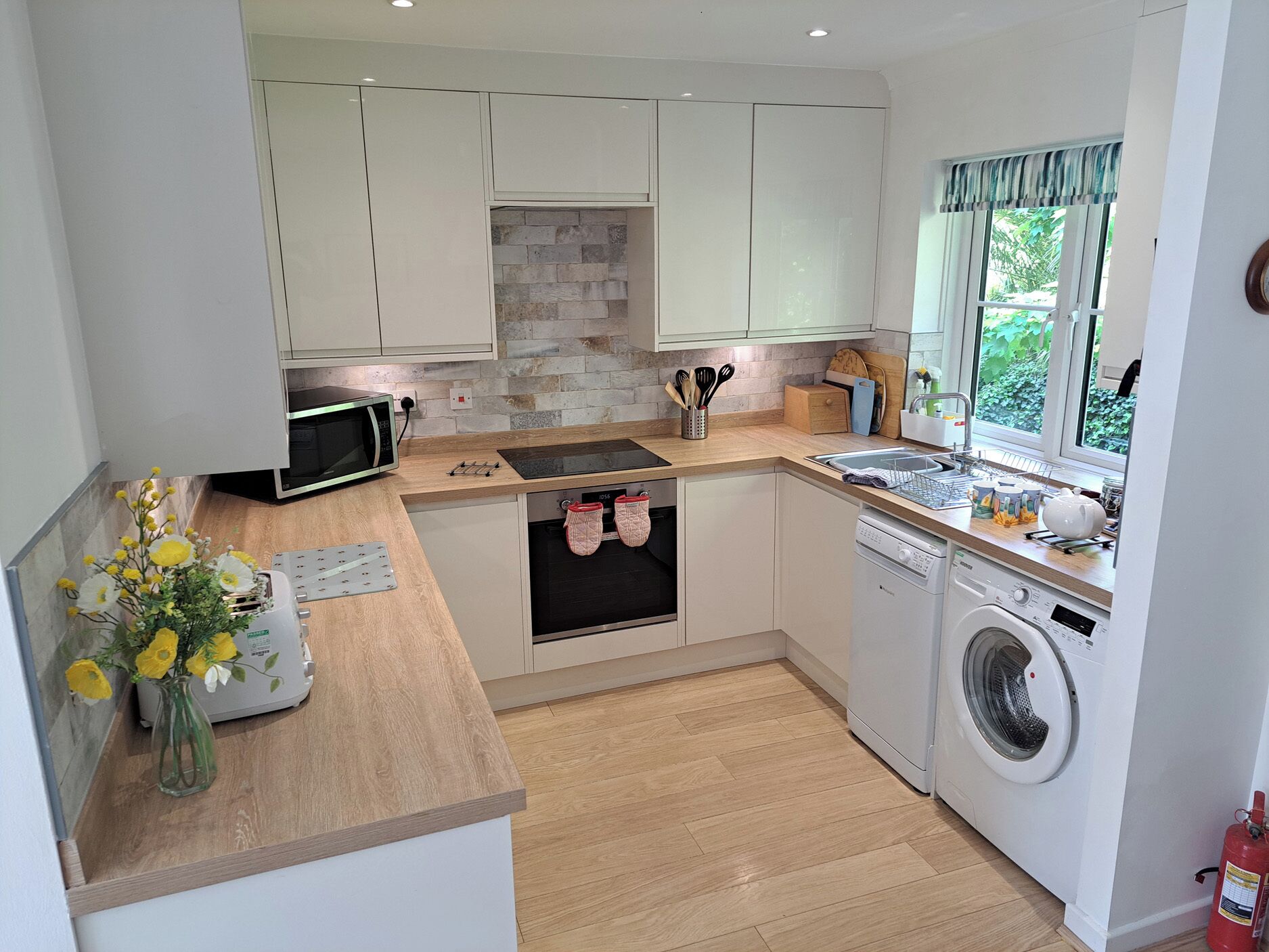 A compact kitchen with a microwave, oven, and washing machine, a small window with a view, and a vase with flowers on the counter.