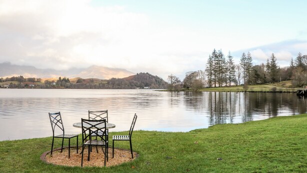 Vista de un lago con una mesa y sillas en una zona cubierta de césped, rodeado de árboles y una cadena montañosa distante.