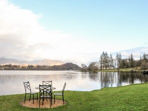 A lakeside view with a table and chairs on a grassy area, surrounded by trees and a distant mountain range.