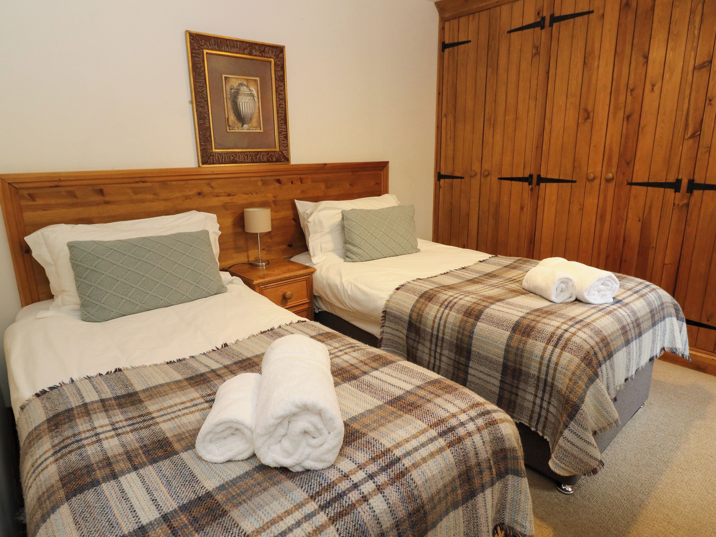 A hotel room with two beds, a wooden wardrobe, a framed picture on the wall, and a bedside table with a lamp.