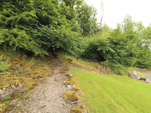A gravel path leading through a forested area with dense green foliage and a grassy area on the right.