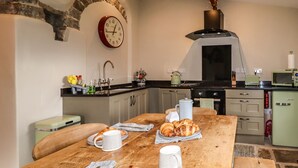 Un tavolo da pranzo in legno con croissant e una tazza, una cucina con elettrodomestici d'epoca e un orologio appeso al muro.