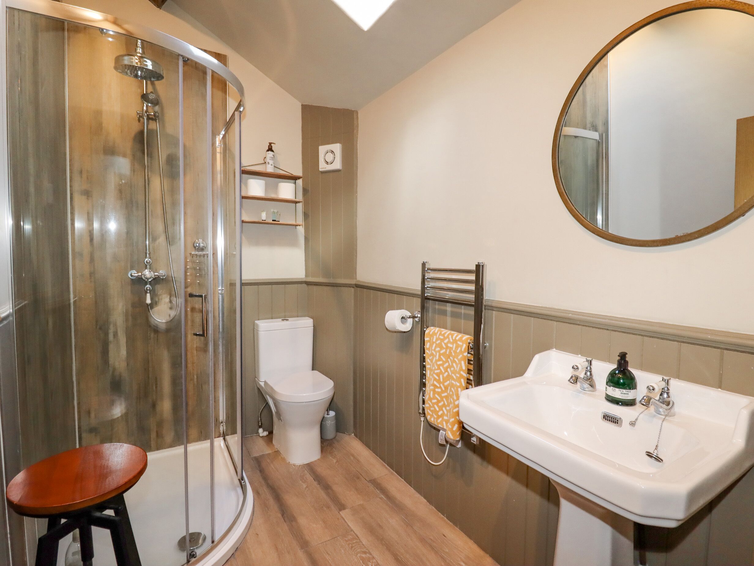 Een badkamer met een douche in glazen cabine, een toilet, een wastafel met spiegel en een houten kruk.