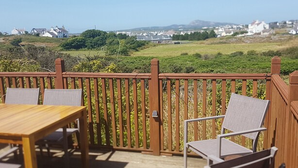 A wooden deck with a table and chairs, overlooking a green landscape with buildings in the distance.