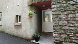 Il s’agit d’un bâtiment en pierre avec une petite fenêtre et une porte. On y voit deux jardinières suspendues et une plante en pot posée sur le sol.