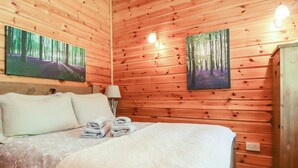 A wooden-paneled bedroom with a bed, two paintings, a nightstand, and a lamp. - Gisburn Forest Lodge (Skipton)