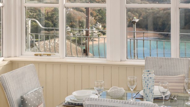 A dining area with a table set for two, overlooking a coastal landscape with hills and a beach. - The Keel Row (Salcombe)