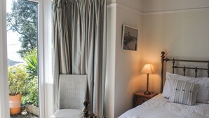 A bedroom with a bed, a chair, a bedside table with a lamp, and a window with curtains. - The Keel Row (Salcombe)