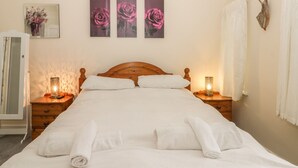 A neatly made bed with white linens and towels, wooden headboard, bedside tables with lamps, and three framed floral paintings on the wall. - Helredale Retreat Whitby (Whitby)