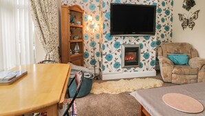 A cozy living room with a fireplace, a television mounted above it, a wooden cabinet, and a comfortable armchair. - Helredale Retreat Whitby (Whitby)