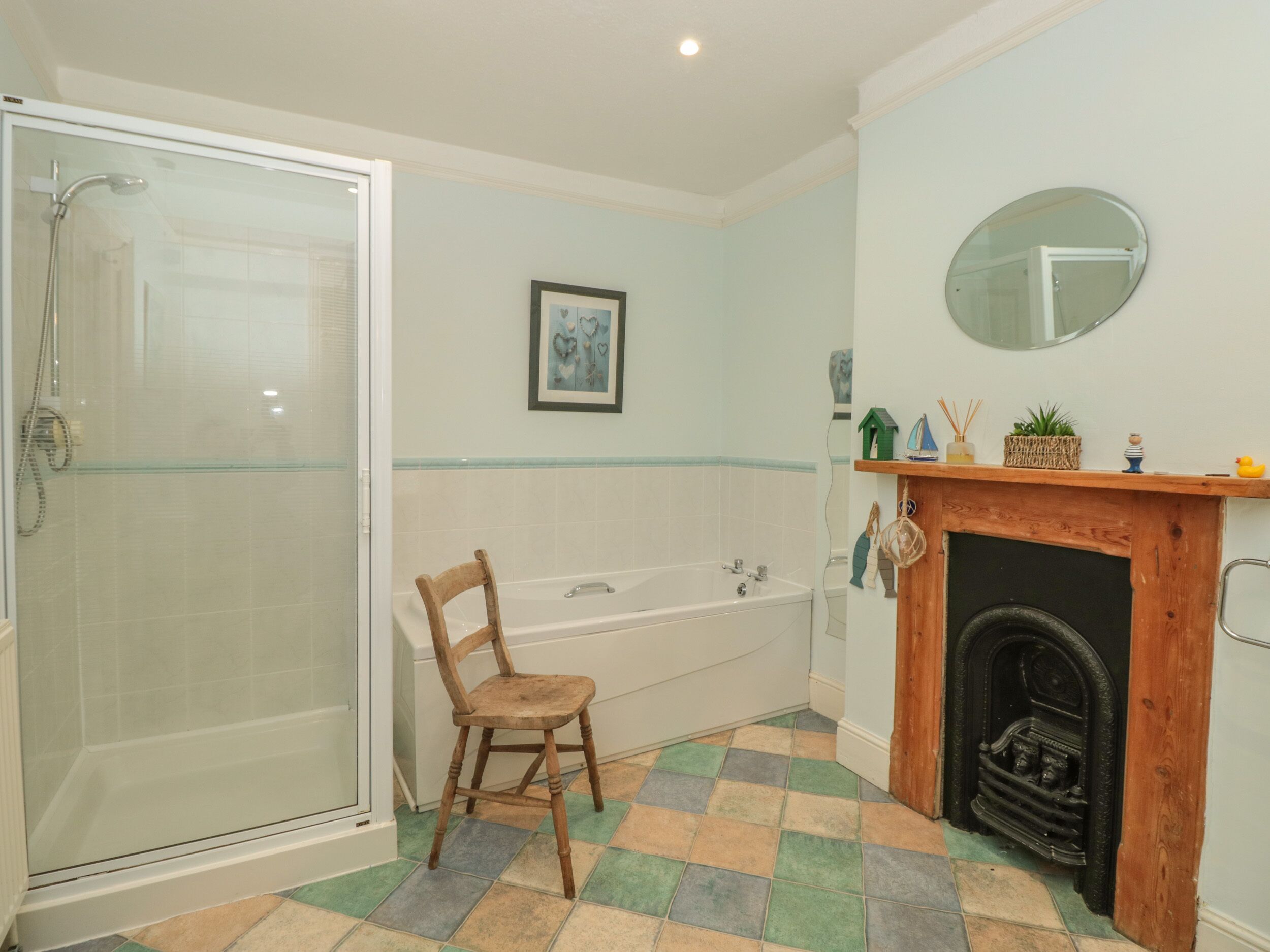 A bathroom with a glass shower enclosure, a wooden chair, a bathtub, a fireplace, and a framed picture on the wall.