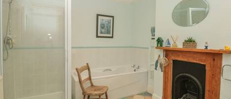 A bathroom with a glass shower enclosure, a wooden chair, a bathtub, a fireplace, and a framed picture on the wall.