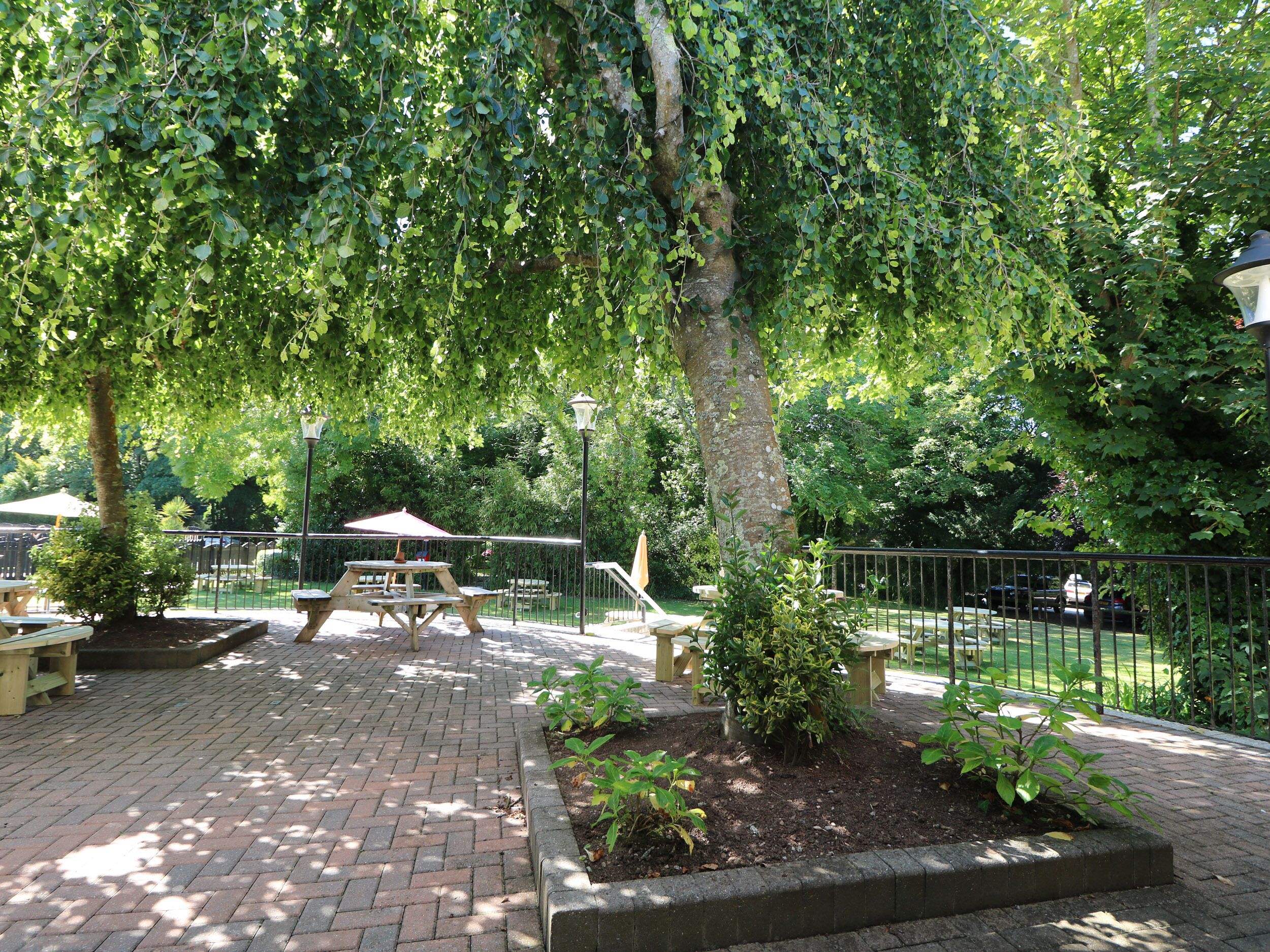 A paved outdoor area with picnic tables and benches, surrounded by trees and a fence.