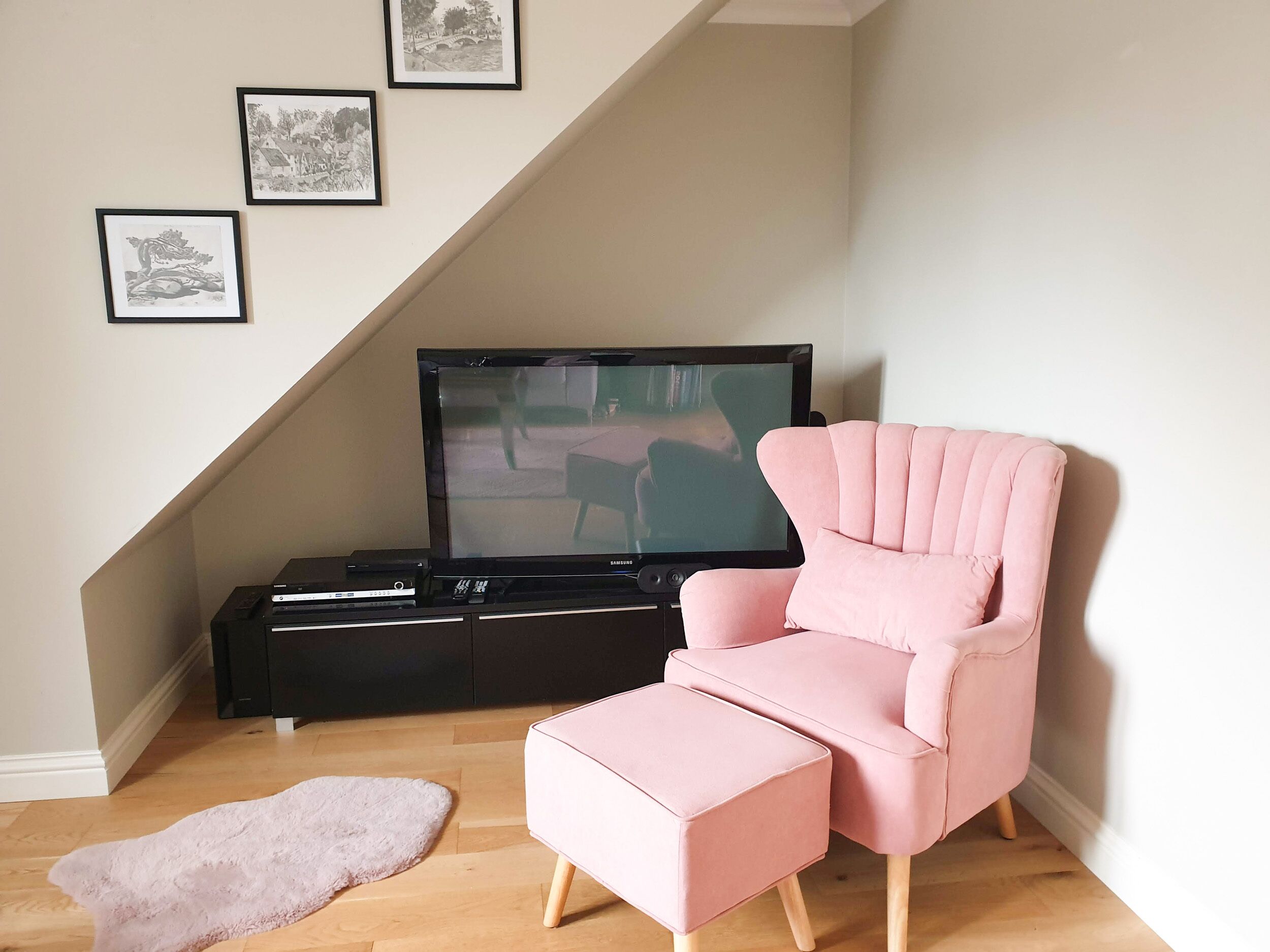 A living room with a pink armchair and ottoman, a black entertainment unit with a TV, and three framed pictures on the wall.