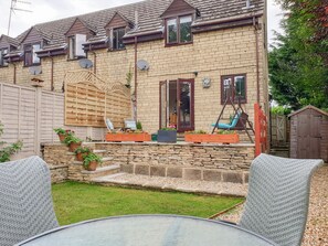 Une maison à deux étages avec une façade en briques, une terrasse en bois et un petit jardin agrémenté de plantes en pot et de meubles d’extérieur.