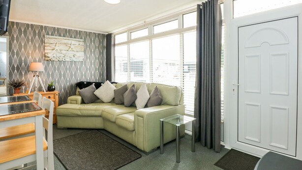 A modern living room with a green sectional sofa, a glass coffee table, and a view through large windows. - 98 Cherry Park (Skegness)