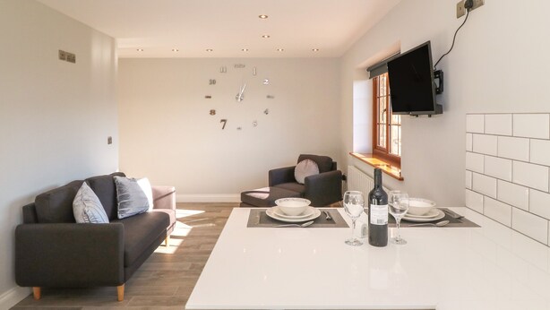 A modern living room with a sofa and armchair, a dining table set with plates, glasses, and a bottle, and a TV mounted on the wall. - Afon Rhos Bach (Caernarfon)