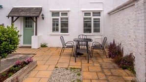 A white building with a green door, a small patio with a table and chairs, and a garden area with flowers. - Martingale (Torquay)