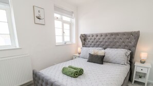 A neatly arranged bedroom with a tufted headboard, a bed with a quilted cover, two bedside lamps, a framed picture on the wall, and a radiator. - Martingale (Torquay)