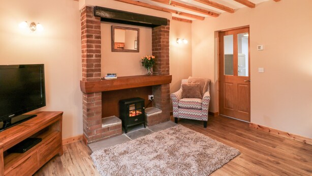 A cozy living room with a brick fireplace, a television on a wooden stand, a patterned rug, and a wooden door. - Barnaby Cottage (Yarm)