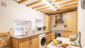 A compact kitchen with wooden cabinets, a built-in washing machine, a microwave, and a stove. There's a breakfast table with croissants and juice. - Barnaby Cottage (Yarm)