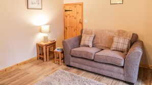 A cozy living room with a wooden ceiling, a brown sofa, a small wooden side table with a lamp, and a rug on the floor. - Barnaby Cottage (Yarm)