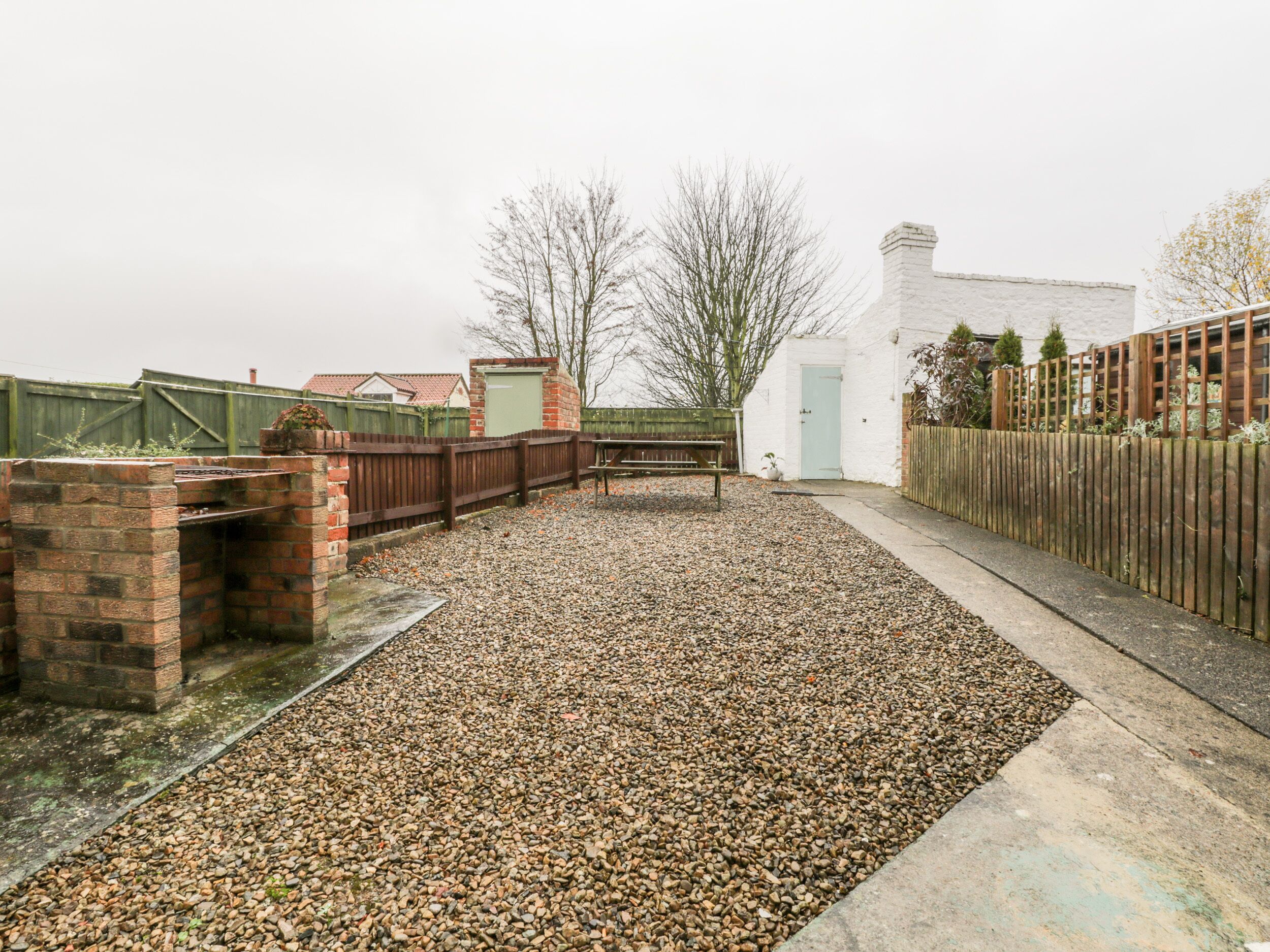 A gravel patio with a brick barbecue area, a wooden fence, and a white building with a door.