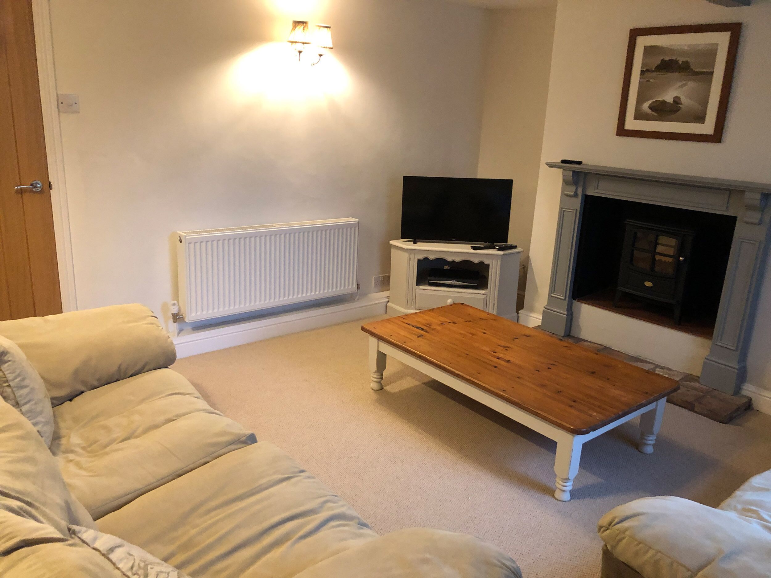 A living room with a beige sofa, a wooden coffee table, a television on a white stand, a white radiator, and a fireplace with a framed picture above it.