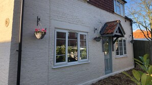 A white brick house with a brown roof, a green door, and two windows with white frames. There are hanging flower baskets on the wall.