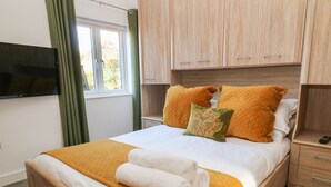 Un letto rifatto con una coperta arancione trapuntata, cuscini bianchi e un cuscino decorativo verde. È presente un comodino in legno con un telecomando sopra.