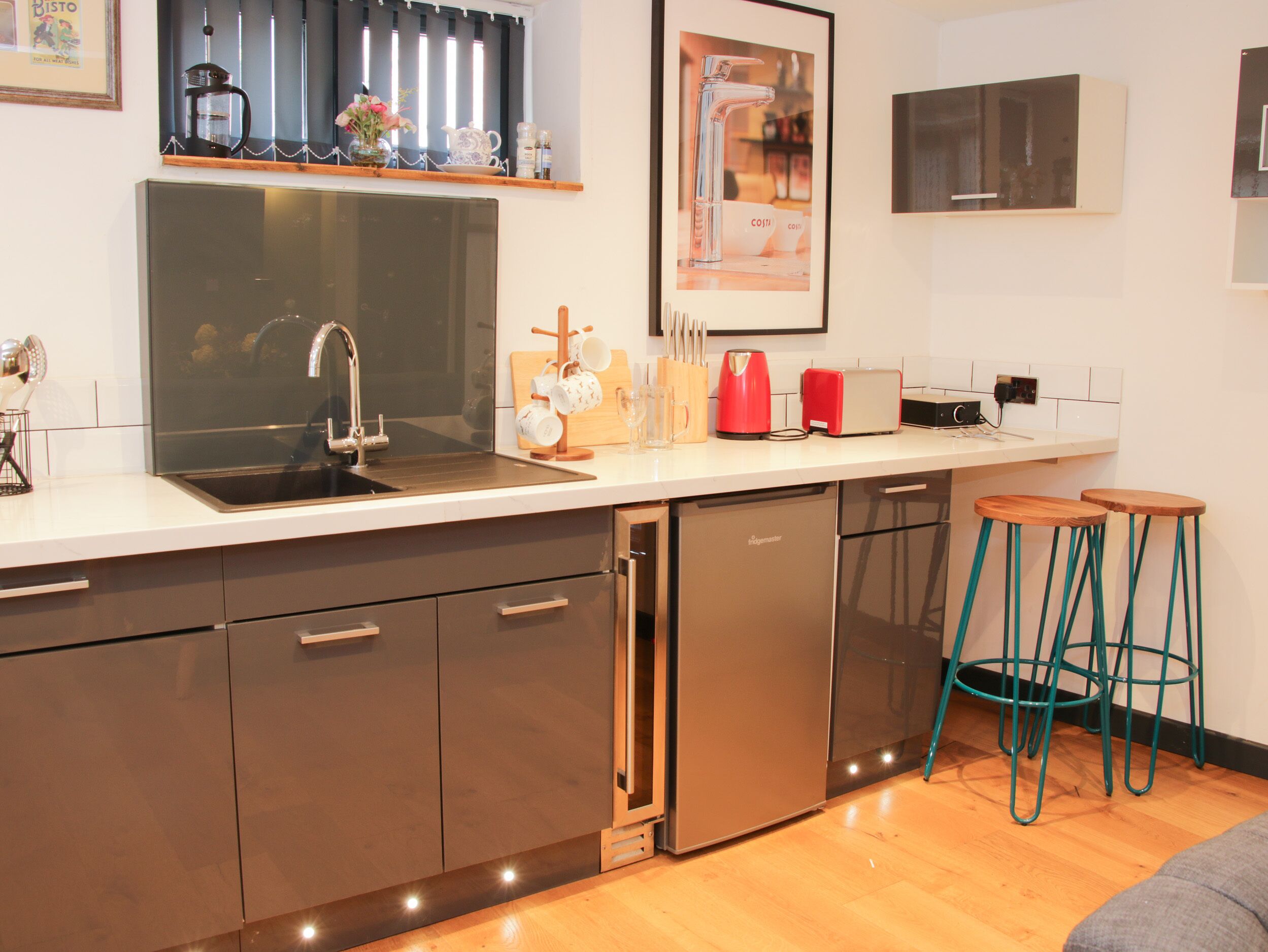 A modern kitchen with a stainless steel sink, a dishwasher, and a coffee machine. There are two stools and a framed picture on the wall.