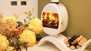 A white wood stove with a glass front displaying a fire, placed on a wooden table next to a vase of yellow flowers. - Bank House Barn (Shrewsbury)