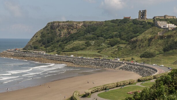 A coastal landscape with a beach, a rocky breakwater, and a historic castle on a hill. - Brinkburn Cottage (Scarborough)