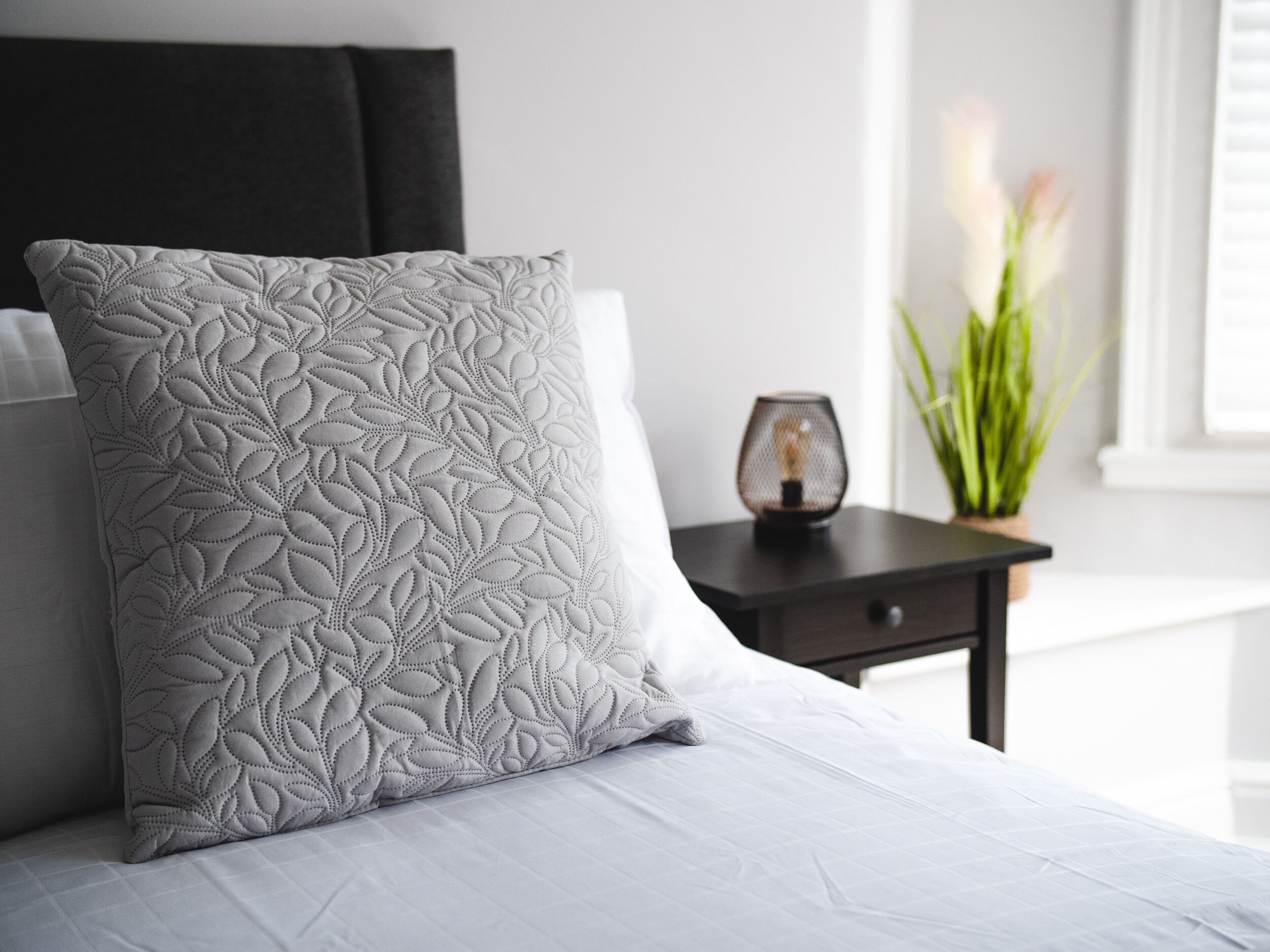 A neatly made bed with a patterned pillow and a bedside table with a lamp and a plant.